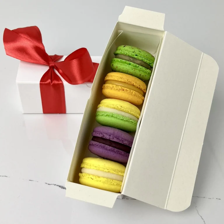 Zdjęcie przedstawia gotowy zestaw do zakupu - makaroniki w białym opakowaniu przewiązane czerwoną kokardką w 5 smakach - zestaw 5 sztuk możliwy do zamówienia przez sklep cukierni Bezownia znajdującej się w Warszawie, ul. F. Joliot-Curie 11