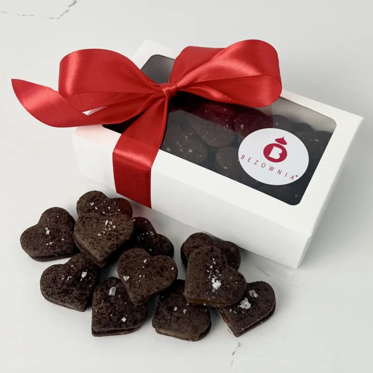 Zdjęcie przedstawia gotowy zestaw do zakupu - serduszka z kajmakiem i płatkami soli w białym opakowaniu przewiązane czerwoną kokardką w 5 smakach - możliwy do zamówienia przez sklep cukierni Bezownia znajdującej się w Warszawie, ul. F. Joliot-Curie 11