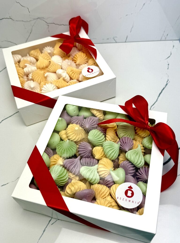 Zdjęcie przedstawia gotowy zestaw do zakupu - kolorowe beziki w białym opakowaniu przewiązane czerwoną kokardką - zestaw możliwy do zamówienia przez sklep cukierni Bezownia znajdującej się w Warszawie, ul. F. Joliot-Curie 11
