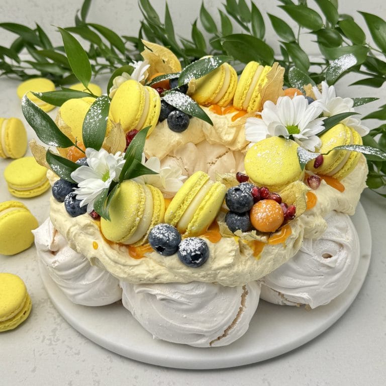 Zdjęcie przedstawia tort bezowy Pavlova z marakują i żółtymi makaronikami, który można zamówić przez sklep cukierni Bezownia znajdującej się w Warszawie, ul. F. Joliot-Curie 11.