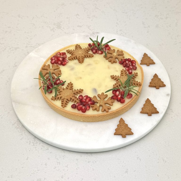 Zdjęcie przedstawia świąteczną tartę z białą czekoladą i czarną porzeczką, udekorowaną pierniczkami. Tarta możliwa jest do zamówienia przez sklep cukierni Bezownia znajdującej się w Warszawie, ul. F. Joliot-Curie 11.