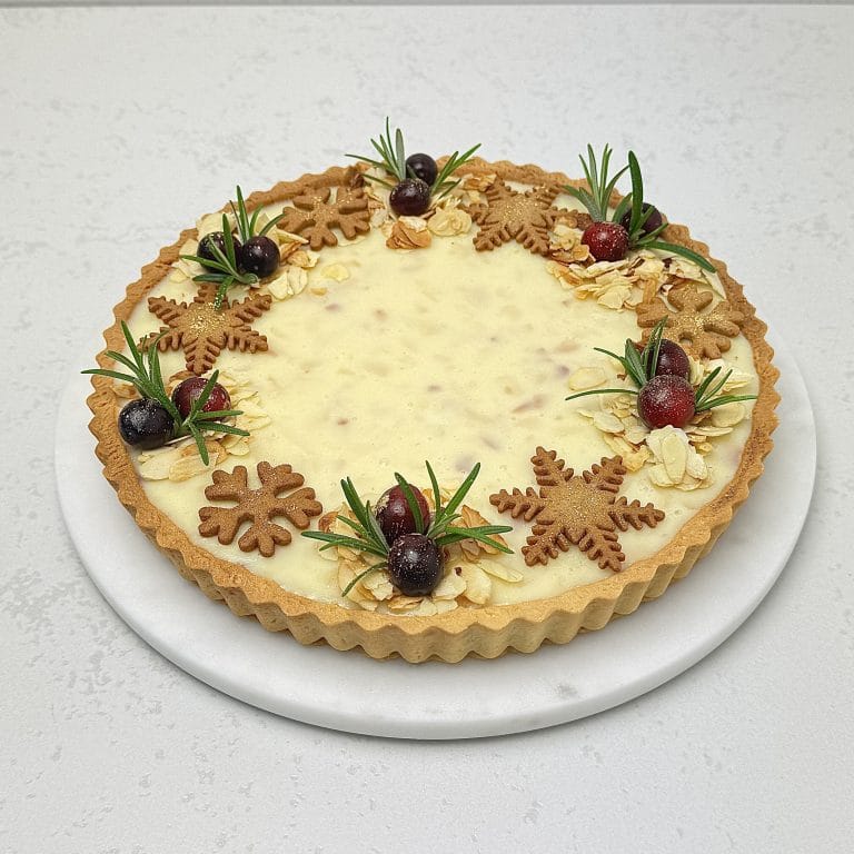 Zdjęcie przedstawia świąteczną tartę z czarną porzeczką i białą czekoladą, udekorowaną pierniczkami. Tarta możliwa jest do zamówienia przez sklep cukierni Bezownia znajdującej się w Warszawie, ul. F. Joliot-Curie 11.