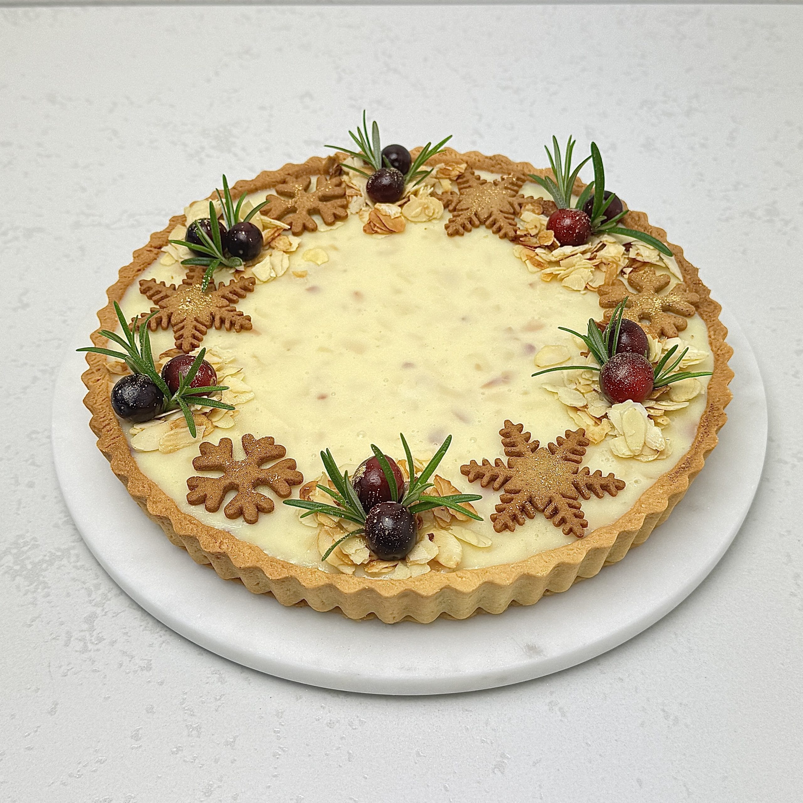 Zdjęcie przedstawia świąteczną tartę z czarną porzeczką i białą czekoladą, udekorowaną pierniczkami. Tarta możliwa jest do zamówienia przez sklep cukierni Bezownia znajdującej się w Warszawie, ul. F. Joliot-Curie 11.